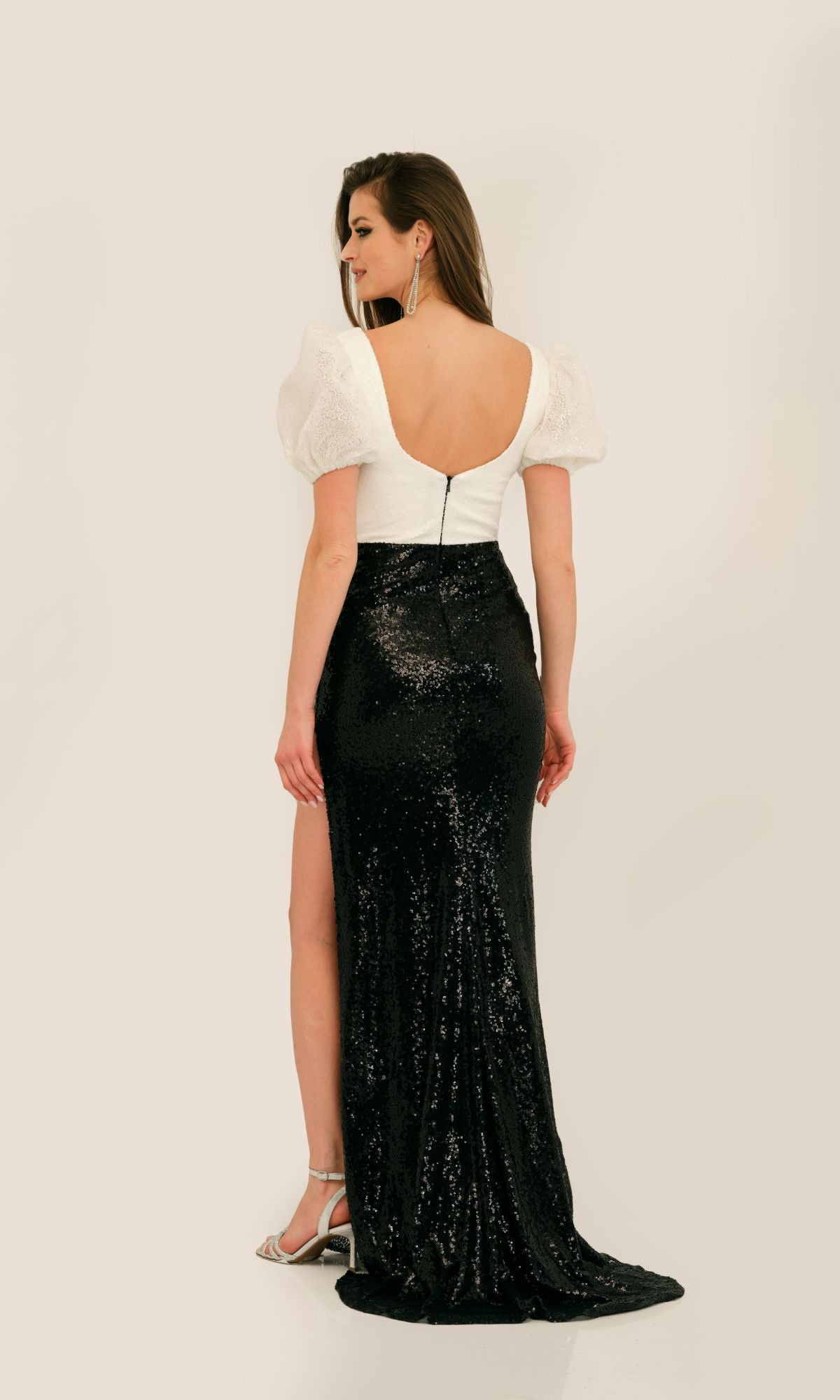 A woman showcases the Long Formal Dress 11328 by Dave and Johnny, a white puff-sleeve, open-back gown paired with a black sequined skirt—an elegant evening look as she stands turned from the camera.